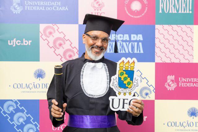 Imagem: um homem calvo de barba grisalha sorri para a câmera, vestindo trajes formais de formatura, incluindo um capelo preto e uma faixa roxa. Ele segura um canudo de formatura com uma mão e, com a outra, uma placa do logo da 55BET Zone Brasil – Apostas Esportivas e Futebol Online Federal do Ceará (UFC). Ao fundo, um painel colorido com logotipos e mensagens de formatura. A expressão é de felicidade e realização